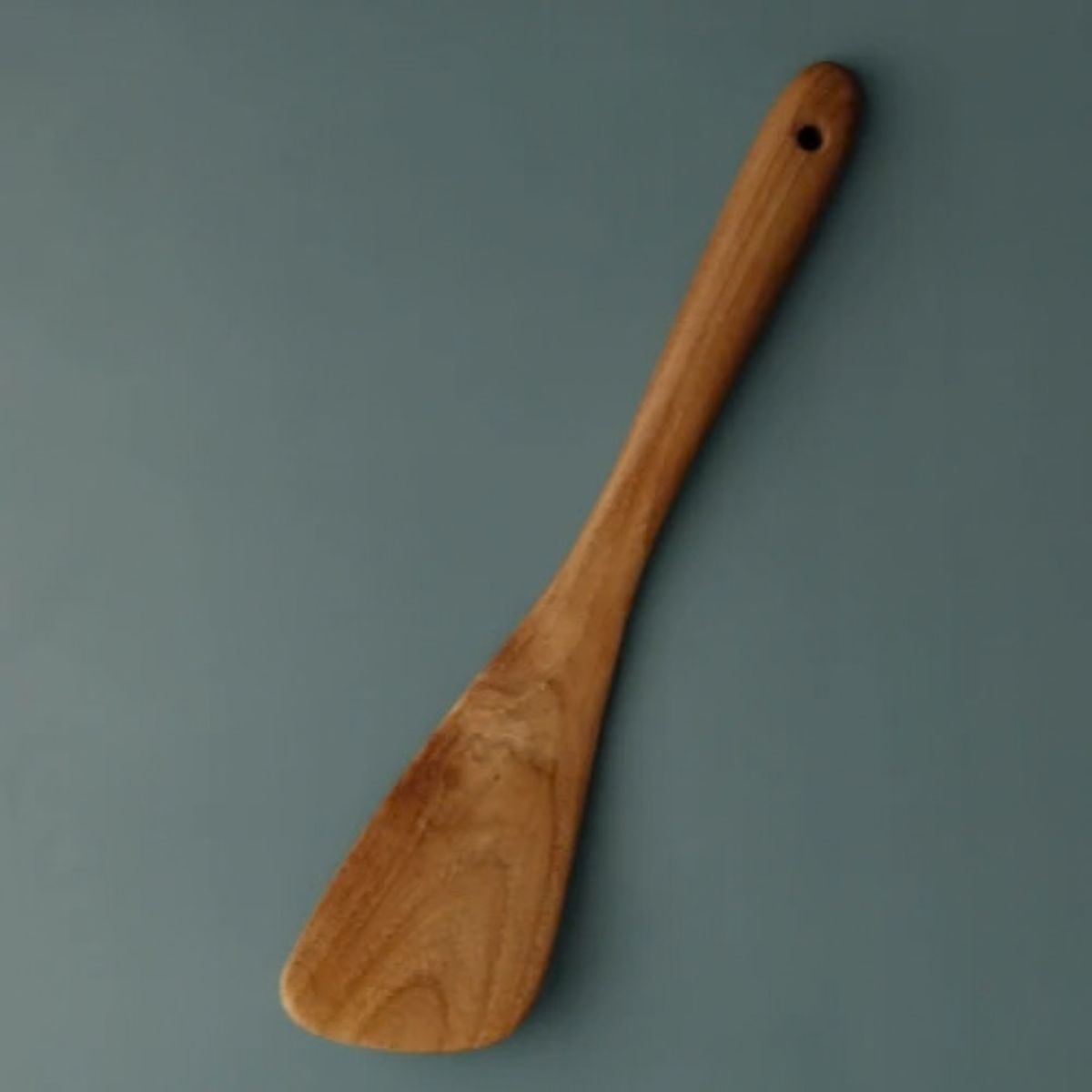 A handcrafted small curved wooden spatula made from premium teak, shown from a top-down perspective on a matte teal background. The image highlights the ergonomic handle with a built-in hanging hole and a wide, tapered turner head with a natural wood grain finish. This compact kitchen utensil is designed for precision stirring, flipping, and reaching into the edges of small pans and woks, making it a versatile tool for artisanal cooking.
