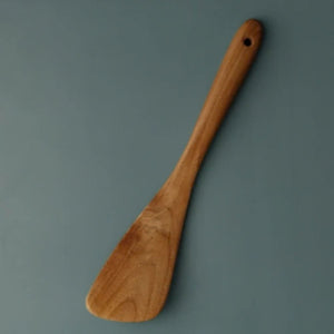 A handcrafted small curved wooden spatula made from premium teak, shown from a top-down perspective on a matte teal background. The image highlights the ergonomic handle with a built-in hanging hole and a wide, tapered turner head with a natural wood grain finish. This compact kitchen utensil is designed for precision stirring, flipping, and reaching into the edges of small pans and woks, making it a versatile tool for artisanal cooking.