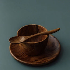 A close-up side-angle photograph of a handcrafted teak wood mealtime set for babies and toddlers. A small, ergonomically curved wooden teaspoon is shown resting horizontally across the rim of a deep, rounded bowl, which sits on a matching circular plate. The set is presented on a smooth, muted grey-blue surface. Carved from single pieces of sustainable, non-toxic teak, each item features a unique, rich natural grain, highlighting its durable and food-safe artisanal construction for early childhood dining.