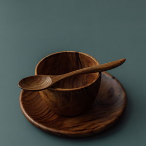 A close-up side-angle photograph of a handcrafted teak wood mealtime set for babies and toddlers. A small, ergonomically curved wooden teaspoon is shown resting horizontally across the rim of a deep, rounded bowl, which sits on a matching circular plate. The set is presented on a smooth, muted grey-blue surface. Carved from single pieces of sustainable, non-toxic teak, each item features a unique, rich natural grain, highlighting its durable and food-safe artisanal construction for early childhood dining.
