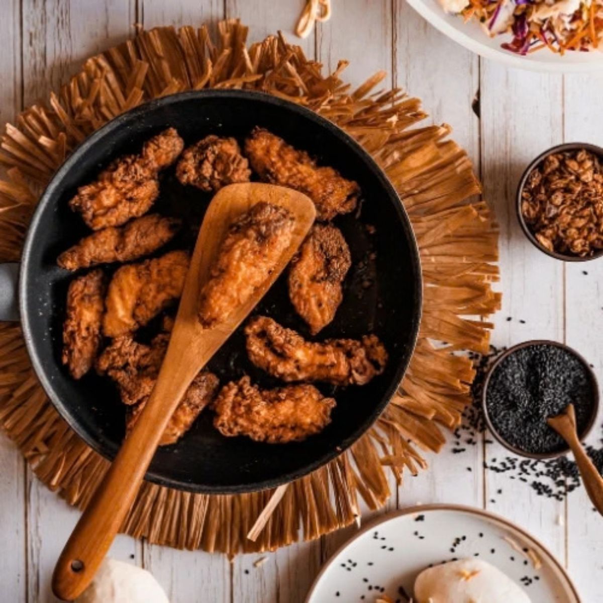 A top-down overhead shot of a handcrafted teak wood cooking spatula being used to flip golden-brown fried chicken in a black non-stick frying pan. The pan sits on a rustic straw mat, surrounded by small bowls of black sesame seeds and crispy toppings. This image demonstrates the wide turner head's effectiveness for heavy-duty frying and highlights the wood's natural heat resistance and non-scratch properties on delicate cookware surfaces.