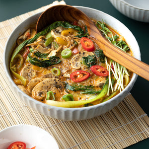 Handcrafted teak wood ladle serving a colorful vegetable soup with mushrooms and chili in a ceramic bowl, placed on a bamboo mat.