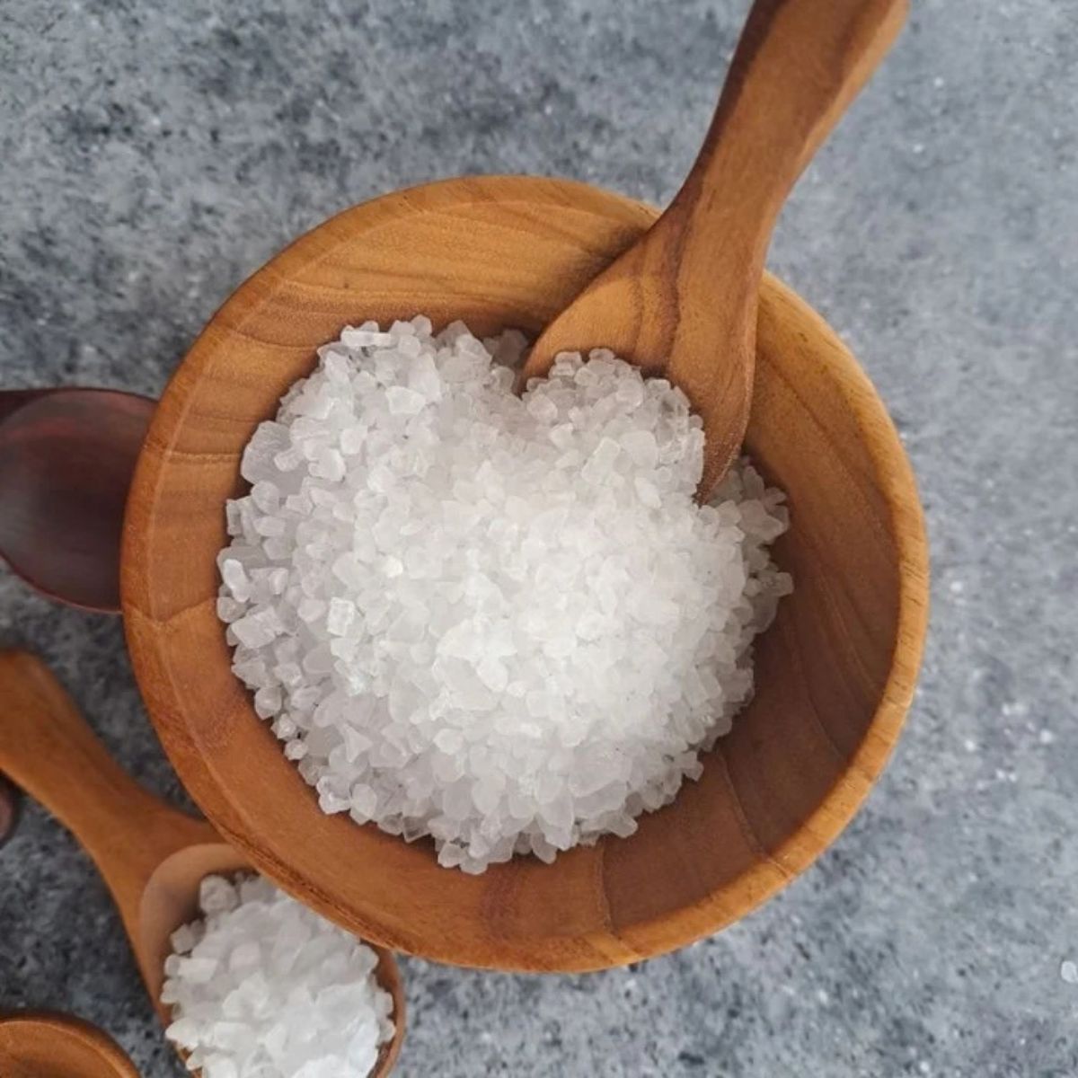 An overhead shot of a handcrafted 8 cm teak wood mini bowl being used as a salt cellar, filled with coarse white sea salt. A small wooden salt spoon rests inside the bowl, while several other artisanal teak spoons in varying natural wood shades are scattered on a grey stone surface around it. The image highlights the bowl's rich grain and its practical use as a sustainable spice or condiment server in a modern kitchen.