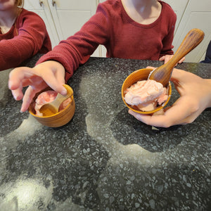 A lifestyle photograph showing two children using handcrafted teak wood mini bowls and small wooden spoons to eat strawberry ice cream. The image is a close-up focusing on their hands and the artisanal bowls on a dark speckled countertop. The natural, warm grain of the teak wood highlights the bowls as safe, non-toxic, and perfectly sized sustainable tableware for kids and toddlers.