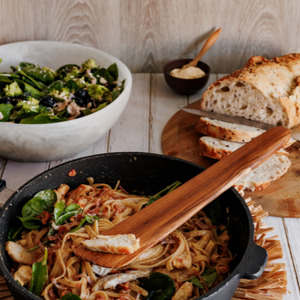 An eye-level lifestyle photograph of a handcrafted teak wood sauté spatula lifting a piece of chicken from a pan of creamy pasta. The spatula is shown in a home dining setting, resting against a black pan on a natural fiber trivet, with a fresh salad and sliced sourdough bread in the background. The warm, golden tones of the teak grain are highlighted by the soft lighting, emphasizing its role as a high-quality, sustainable kitchen tool for both cooking and serving.