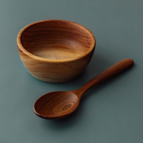 A clean, top-down product photograph of a handcrafted 11 cm teak wood toddler bowl and its matching rounded wooden spoon, arranged symmetrically on a smooth, muted grey-blue surface. The image showcases the rich, swirling natural grain of the sustainable teak, highlighting the set's smooth, non-toxic, and food-safe finish. Both the bowl and spoon are carved from single
