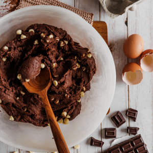 A top-down lifestyle photograph of "My First Big Spoon" being used to mix thick chocolate cookie dough with white chocolate chips in a large white bowl. The scene is set on a rustic white wooden table surrounded by baking ingredients, including fresh eggs, cocoa powder, and chocolate bars. The handcrafted teak wood spoon's wide head and sturdy handle are shown effectively handling the heavy dough, highlighting its durability and non-toxic, food-safe nature for children participating in kitchen activities.
