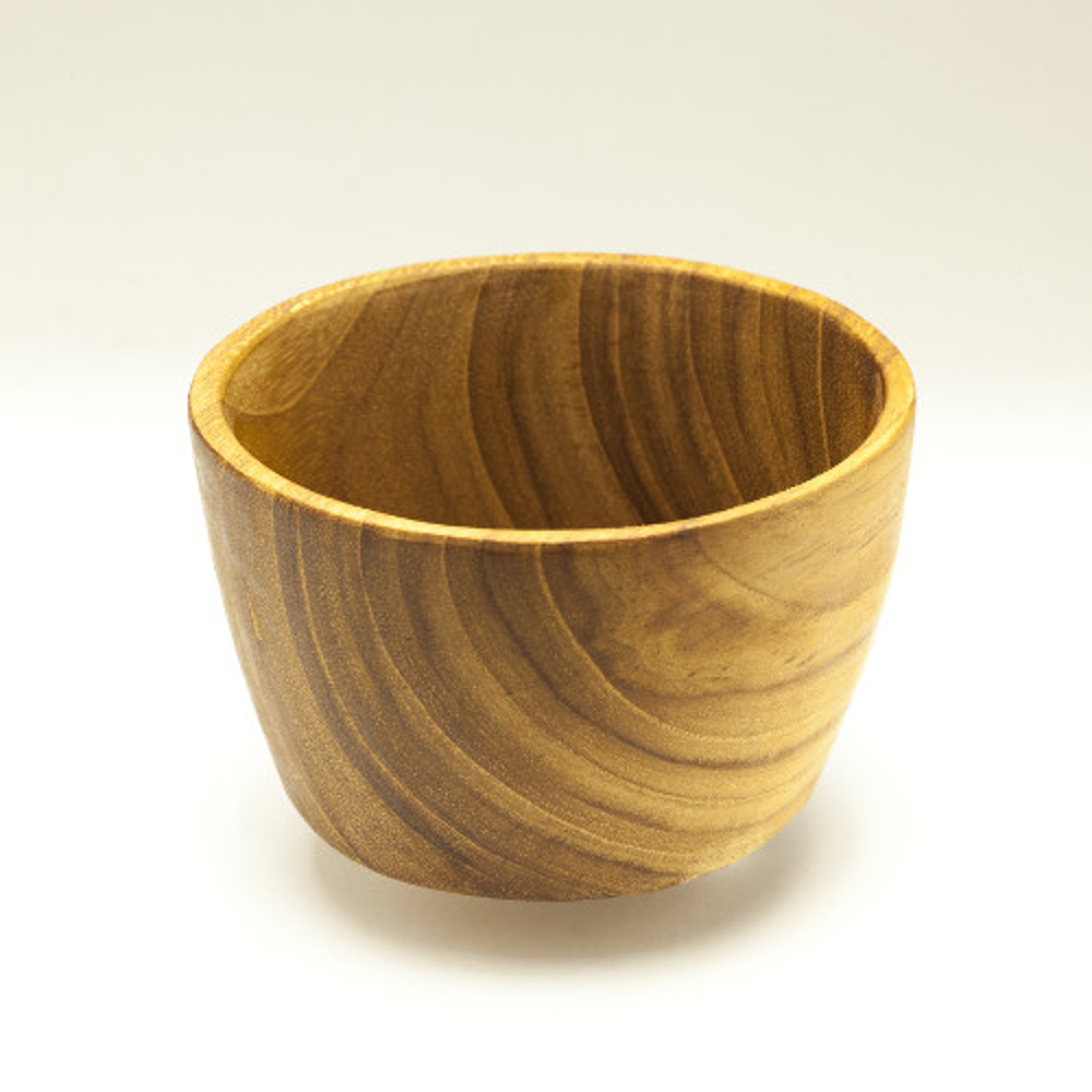 A top-down lifestyle photograph of a children’s mealtime set featuring a 13 cm handcrafted teak wood bowl and a matching rounded wooden spoon. The bowl and spoon are nested on a larger flat teak plate, all set against a smooth, muted grey-blue background. The image showcases the unique, swirling natural wood grain of the sustainable teak, highlighting the set's durable, non-toxic, and plastic-free construction designed for toddlers.