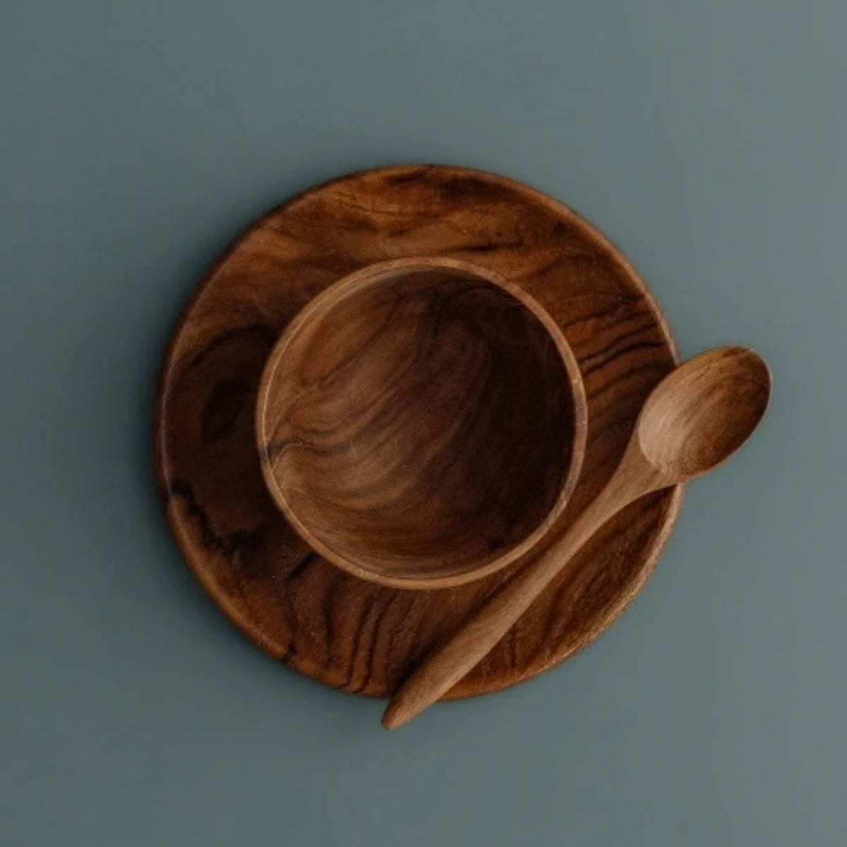 An overhead "flat lay" photograph of the Safer Mealtime Set for babies and toddlers, featuring a handcrafted teak wood bowl nested in the center of a matching teak plate. A small wooden teaspoon is placed vertically to the right of the bowl. The entire set is presented on a smooth, muted grey-blue background, highlighting the rich, swirling natural grain of the sustainable wood. Carved from single pieces of food-safe teak, this durable and non-toxic tableware set offers a healthy, plastic-free alternative