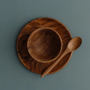 An overhead "flat lay" photograph of the Safer Mealtime Set for babies and toddlers, featuring a handcrafted teak wood bowl nested in the center of a matching teak plate. A small wooden teaspoon is placed vertically to the right of the bowl. The entire set is presented on a smooth, muted grey-blue background, highlighting the rich, swirling natural grain of the sustainable wood. Carved from single pieces of food-safe teak, this durable and non-toxic tableware set offers a healthy, plastic-free alternative