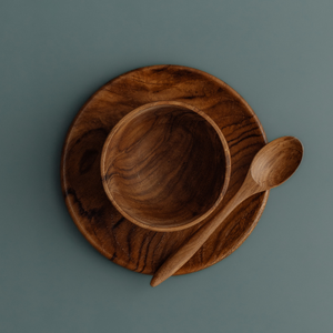 An overhead "flat lay" photograph of the Safer Mealtime Set for babies and toddlers, featuring a handcrafted teak wood bowl nested in the center of a matching teak plate. A small wooden teaspoon is placed vertically to the right of the bowl. The entire set is presented on a smooth, muted grey-blue background, highlighting the rich, swirling natural grain of the sustainable wood. Carved from single pieces of food-safe teak, this durable and non-toxic tableware set offers a healthy, plastic-free alternative f