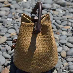 A hand-woven Krajood sea grass bucket bag in a natural golden-tan tone, featuring a sturdy dark brown leather handle and wooden bead accents, resting on smooth grey river stones.