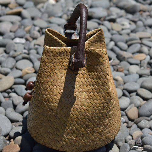 A hand-woven Krajood sea grass bucket bag in a natural golden-tan tone, featuring a sturdy dark brown leather handle and wooden bead accents, resting on smooth grey river stones.