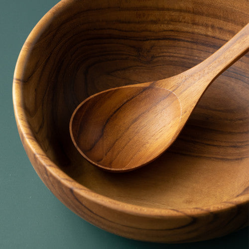 A detailed close-up of a handcrafted teak wood soup spoon resting inside a matching teak wood bowl. The image highlights the intricate, swirling natural wood grain patterns and the smooth, polished surface of both pieces against a deep forest green background.