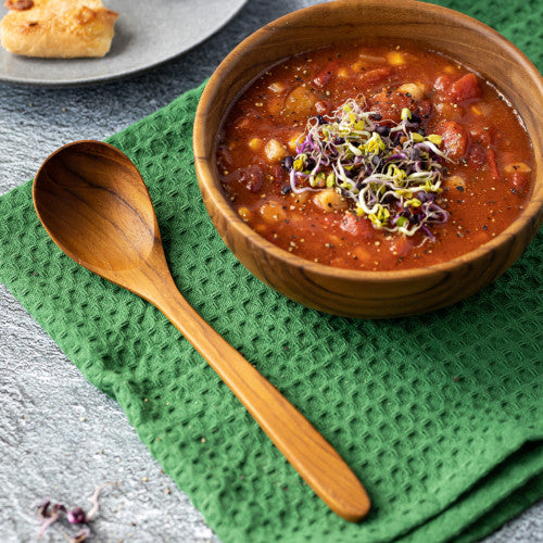 A lifestyle shot of a handcrafted 18cm teak wood soup spoon resting on a green waffle-weave napkin next to a matching wooden bowl filled with hearty tomato and vegetable soup. The scene includes a side plate with bread, highlighting the natural grain of the teak against a rustic grey tabletop.