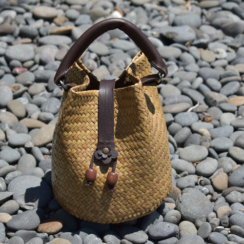 A hand-woven Krajood sea grass bucket bag in a warm tan color, featuring a dark brown leather top handle and decorative wooden bead drawstring pulls, displayed on a bed of natural smooth river stones.