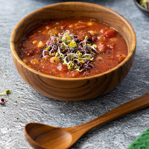 A close-up lifestyle image of a handcrafted teak wood soup spoon positioned in front of a matching wooden bowl filled with tomato and bean soup topped with fresh sprouts. The photo highlights the warm, natural grain of the teak wood against a textured grey stone surface.
