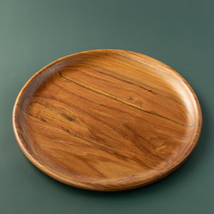 A handcrafted small teak wood plate, 35 cm in diameter, isolated on a smooth dark green background. The image showcases the rich, golden-brown natural wood grain and the polished, food-safe finish of the circular platter. This versatile small plate is ideally sized for serving side dishes, tasting portions, appetizers, or individual slices of cake and cheesecake in a sustainable, artisanal kitchen setting.