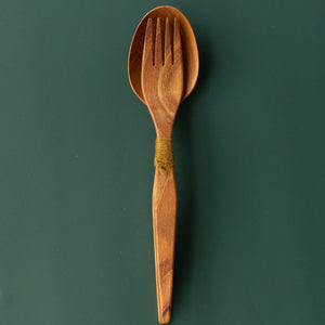Handcrafted teak wood salad server set with a fork nested in a spoon tied with green yarn, shown from a top-down view on a dark green surface.