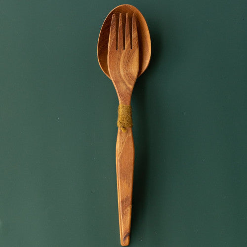 Handcrafted teak wood salad server set with a fork nested in a spoon tied with green yarn, shown from a top-down view on a dark green surface.