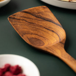 A high-detail close-up of the wide, solid head of a handcrafted medium teak wood spatula. The image, set against a dark forest green background, showcases the rich, swirling natural grain and deep golden-brown tones of the premium teak wood. This polished, non-scratch surface is designed for flipping and serving delicate foods, emphasizing the artisanal quality and durable construction of the kitchen utensil.