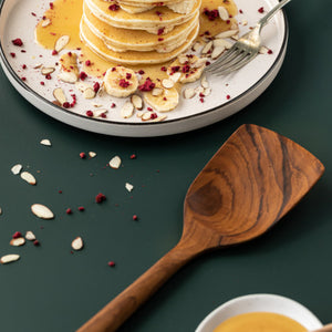 A lifestyle cooking photograph featuring a handcrafted teak wood spatula resting on a dark forest green surface next to a stack of fresh pancakes. The pancakes are served on a white ceramic plate and garnished with sliced bananas, almonds, and syrup. The image highlights the wide, solid turner head and the rich, dark swirling wood grain of the teak, demonstrating its use as a gentle, non-scratch tool for flipping delicate breakfast items and serving in a modern kitchen setting.